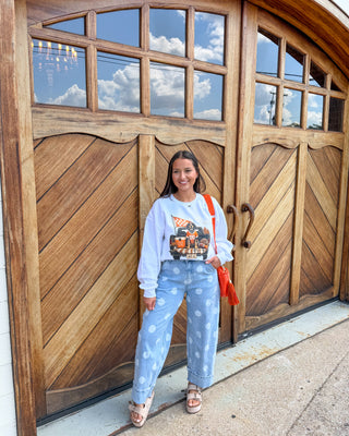 A woman stands in front of large wooden garage-style doors wearing a light blue sweatshirt with a graphic print, matching light blue patterned pants, and sandals, while carrying a bright orange shoulder bag.
