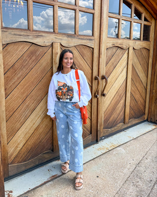 A woman stands in front of large rustic wooden garage doors, wearing a white sweatshirt with a colorful graphic print, light blue polka-dot sweatpants, and sandals, and carrying a bright orange shoulder bag.