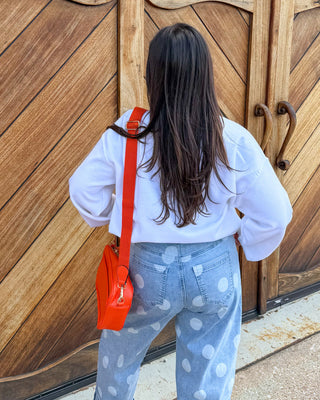 A woman viewed from behind stands in front of large rustic wooden garage-style doors, wearing a white long-sleeve top, light blue jeans with white polka dots, and carrying a bright orange shoulder bag.