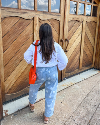 A woman viewed from behind stands in front of large rustic wooden garage-style doors, wearing a white long-sleeve top, light blue jeans with white polka dots, and carrying a bright orange shoulder bag.