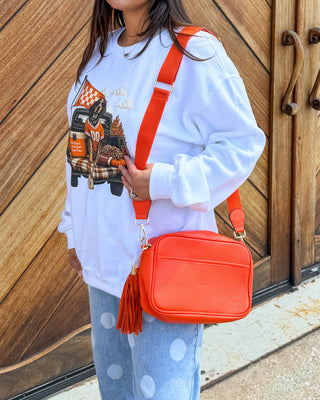 A woman stands in front of large wooden garage-style doors wearing a white graphic sweatshirt, light blue patterned pants, and sandals, while carrying a bright orange crossbody bag.