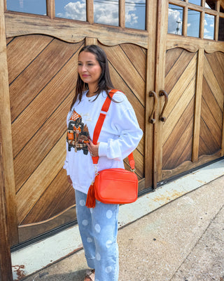 A woman stands in front of large wooden garage-style doors wearing a white graphic sweatshirt, light blue patterned pants, and sandals, while carrying a bright orange crossbody bag.