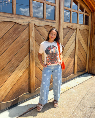 A woman stands in front of a large wooden garage door wearing a white graphic t-shirt with a dog print, light blue wide-leg jeans with white polka dots, and sandals, carrying a red shoulder bag.