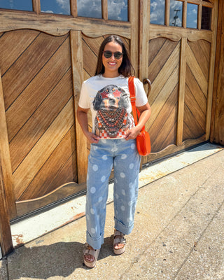 A woman stands in front of large wooden garage doors wearing a white graphic t-shirt, light blue wide-leg jeans with white polka dots, brown sandals, and carrying an orange shoulder bag.