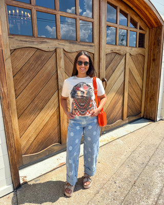 A woman stands in front of large wooden garage doors wearing a white graphic t-shirt, light blue wide-leg jeans with white polka dots, brown sandals, and carrying an orange shoulder bag.