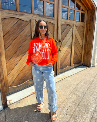 A woman wearing a bright orange sweatshirt with white graphic text, light blue patterned jeans, and brown sandals stands in front of a large wooden garage door, holding a round orange item.