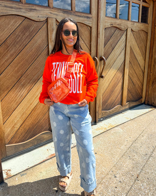A woman wearing a bright orange sweatshirt with white graphic text, light blue patterned jeans, and brown sandals stands in front of a large wooden garage door, holding a round orange item.
