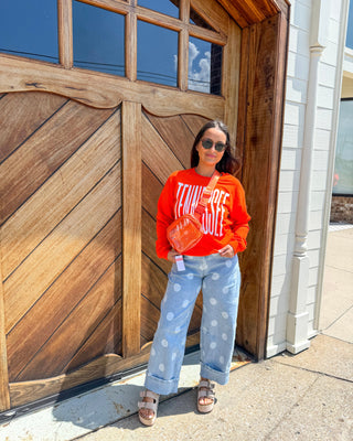 A woman stands in front of a large wooden garage door wearing a bright orange sweatshirt with white text, light blue wide-leg jeans with white polka dots, and brown sandals. She is holding a small orange bag.