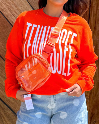 A woman stands in front of a large wooden garage door wearing a bright orange sweatshirt with white text, light blue wide-leg jeans with white polka dots, and brown sandals. She is holding a small orange bag.