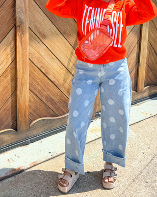 A woman posing outdoors in front of a large wooden garage door, wearing a bright orange sweatshirt with 'TENNESSEE' printed on it, light blue polka dot jeans, and sunglasses.