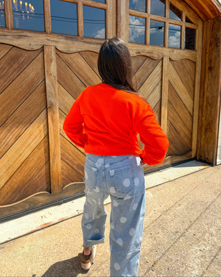 A person viewed from behind wearing a bright orange long-sleeve crop top and light blue jeans with white polka dots, standing in front of a large wooden garage door.