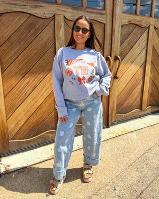 A woman wearing a light blue graphic sweatshirt and light blue polka dot jeans stands in front of a large wooden garage door. The sweatshirt features a graphic with the words 'The Night' and 'TX' with orange and red details.
