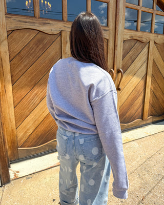 A person viewed from behind wearing a light heather gray sweatshirt and light blue jeans with white polka dots, standing in front of a large wooden door.