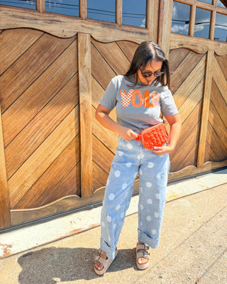 A woman stands outdoors in front of a large wooden garage door, wearing a gray graphic T-shirt with orange lettering, light blue wide-leg polka dot jeans, and brown sandals, while holding a bright orange textured clutch.