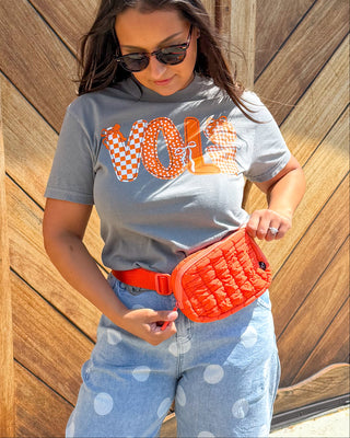 A woman stands outdoors in front of a large wooden garage door, wearing a gray graphic T-shirt with orange lettering, light blue polka-dot jeans, a bright orange fanny pack, and brown sandals.