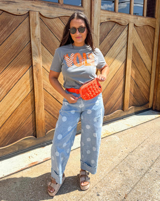A woman stands outdoors in front of a large wooden garage door, wearing a gray graphic T-shirt, light blue polka-dot jeans, a bright orange woven belt bag, and brown sandals.