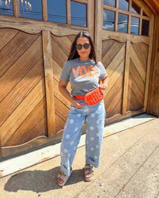 A woman stands outdoors in front of a large wooden garage door, wearing a gray graphic T-shirt, light blue polka-dot jeans, a bright orange woven belt bag, and brown sandals.