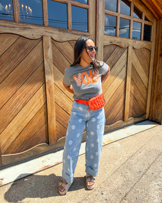 A woman stands outdoors in front of a large wooden garage door, wearing a gray graphic t-shirt with orange lettering, light blue polka dot jeans, a bright orange quilted fanny pack, and brown sandals.