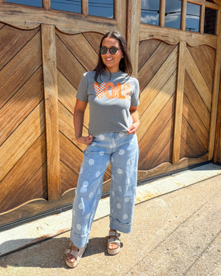 A woman stands outdoors in front of a large wooden garage door wearing a gray graphic t-shirt with orange lettering, light blue wide-leg jeans with white polka dots, and brown sandals.