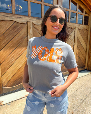 A woman wearing a gray t-shirt with a bold orange and white 'VOLE' graphic, paired with light blue polka-dot jeans, standing in front of a rustic wooden garage door.