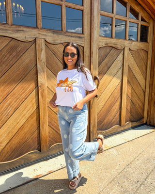 A woman wearing a white graphic t-shirt with a leopard print, light blue jeans, and brown sandals is posing in front of a large wooden garage door.