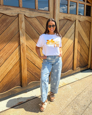 A woman stands in front of a large wooden garage door wearing a white graphic t-shirt with a yellow and orange 'SUNSHINE VILLAGE' design, light-wash distressed jeans, and brown sandals.
