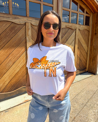 A woman wearing a white graphic t-shirt with an orange and brown Tennessee 'TN' map design, paired with light blue jeans, standing in front of a large wooden garage door.