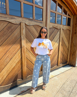 A woman stands in front of a large wooden garage door wearing a white graphic t-shirt with a yellow giraffe print, light blue wide-leg jeans with white polka dots, and brown sandals.