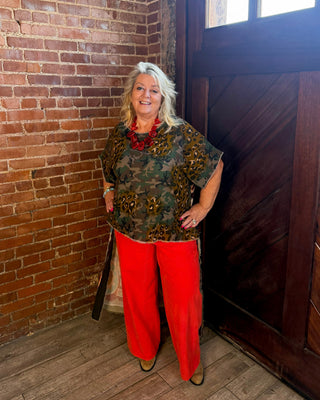 A woman stands in a rustic room with a brick wall and a dark wooden door, wearing a green and brown patterned short-sleeve top, bright red wide-leg pants, and a red necklace.
