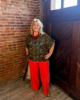 A woman with white hair stands in a rustic room with a brick wall and a dark wooden door, wearing a green and brown patterned short-sleeve top, bright red wide-leg pants, and a red necklace.
