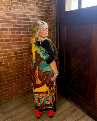A woman stands in a corner with a brick wall and a dark wooden door, wearing a long, colorful graphic print wrap or dress over a black top and red tights.