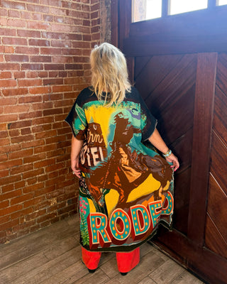 A person stands with their back to the camera in a rustic indoor setting with a brick wall and wooden door, wearing a black graphic t-shirt with a large colorful western-themed print that reads 'Rodeo' and 'Don't Tread on Me', paired with bright red pants.