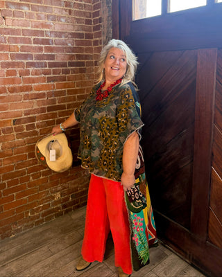 An older woman with gray hair stands in a rustic room with a brick wall and dark wooden door, wearing a patterned green top, bright red pants, and holding a straw hat.