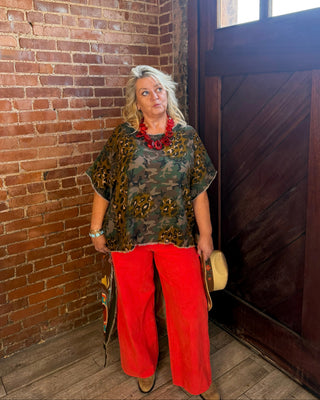 A woman stands in a rustic indoor setting with a brick wall and a dark wooden door, wearing a short-sleeve camouflage-patterned top with yellow floral details, bright red wide-leg pants, and a chunky red beaded necklace.