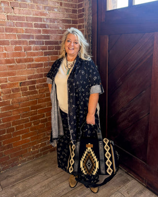 A woman with gray hair stands in a rustic room with a brick wall and wooden door, wearing a black patterned kimono-style cardigan over a cream top and holding a black tote bag with a geometric pattern.