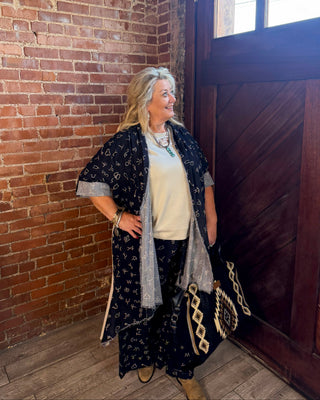 A woman stands in a rustic room with a brick wall and dark wooden door, wearing a bohemian-style outfit featuring a navy blue patterned kimono over a cream top and matching pants, holding a black and beige patterned handbag.