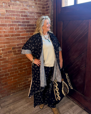 A woman stands in a rustic room with a brick wall and dark wooden door, wearing a bohemian-style outfit featuring a navy blue patterned kimono over a cream top and matching pants, holding a black and beige patterned handbag.
