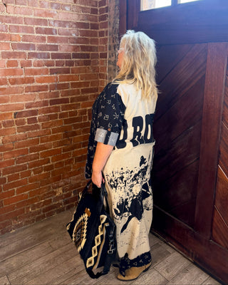 A woman stands in a corner with a red brick wall and a dark wooden door, wearing a cream and black graphic print kimono-style top and holding a black patterned tote bag.