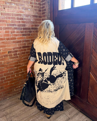 A woman viewed from behind wearing a cream and black long-sleeve cardigan with 'RODEO!' and a cowboy graphic on the back, standing in a rustic room with a brick wall and wooden door.