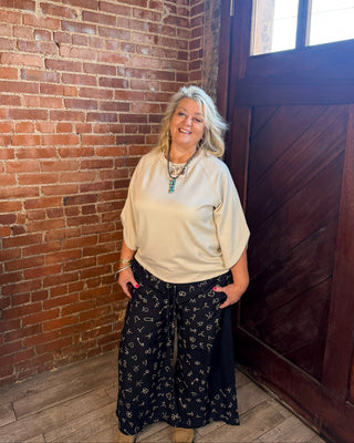 A woman with blonde hair stands in a rustic room with a red brick wall and a dark wooden door, wearing a beige short-sleeve top and black wide-leg pants with a small white floral pattern.