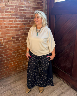 A woman with blonde hair stands in a rustic room with a brick wall and a dark wooden door, wearing a cream-colored top and black wide-leg pants with a small floral pattern.