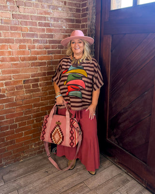 A woman stands in a rustic room with a brick wall and wooden door, wearing a pink hat, a brown striped shirt with a colorful graphic, a long dusty rose skirt, and holding a pink patterned handbag.