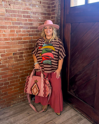 A woman stands in a rustic room with a brick wall and wooden door, wearing a pink hat, a brown striped shirt with a colorful graphic, a long dusty rose skirt, and holding a pink patterned handbag.