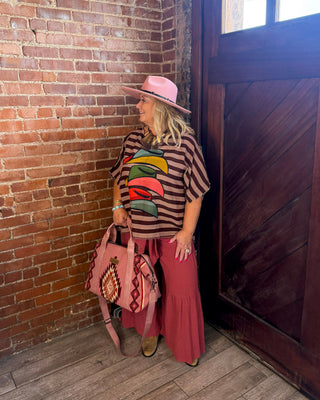 A woman stands in a rustic room with a brick wall and wooden door, wearing a pink hat, a brown and black striped shirt with a colorful graphic, a long pink skirt, and holding a pink patterned handbag.