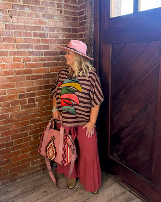 A woman stands in a rustic room with a brick wall and wooden door, wearing a pink hat, a brown and black striped shirt with a colorful graphic, a long pink skirt, and holding a pink patterned handbag.
