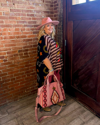 A woman wearing a pink hat and a black poncho with colorful geometric patterns stands in a rustic room with a brick wall and wooden door, holding a pink handbag with a Southwestern-style geometric pattern.