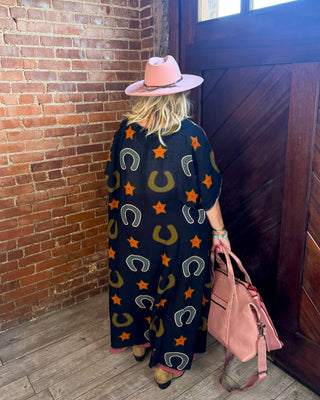 A woman viewed from behind wearing a black poncho with a western-themed print of orange stars, olive horseshoes, and white buckles, paired with a pink hat and holding a pink handbag in a rustic brick and wood doorway.