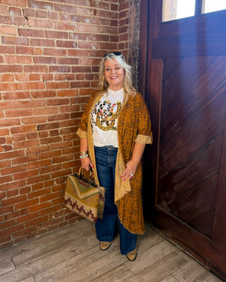A woman stands in a rustic room with a brick wall and wooden door, wearing a white graphic t-shirt, blue jeans, a brown patterned cardigan, and holding a matching patterned handbag.