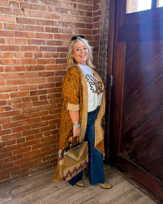 A woman stands in a rustic room with a red brick wall and a dark wooden door, wearing a mustard yellow patterned cardigan over a white graphic tee, blue jeans, and tan boots, while holding a patterned handbag.