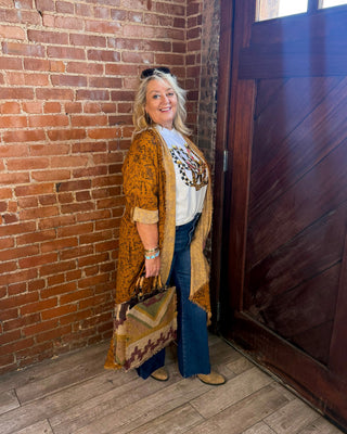 A woman stands in a rustic room with a red brick wall and a dark wooden door, wearing a mustard yellow patterned cardigan over a white graphic tee, blue jeans, and tan boots, while holding a patterned handbag.